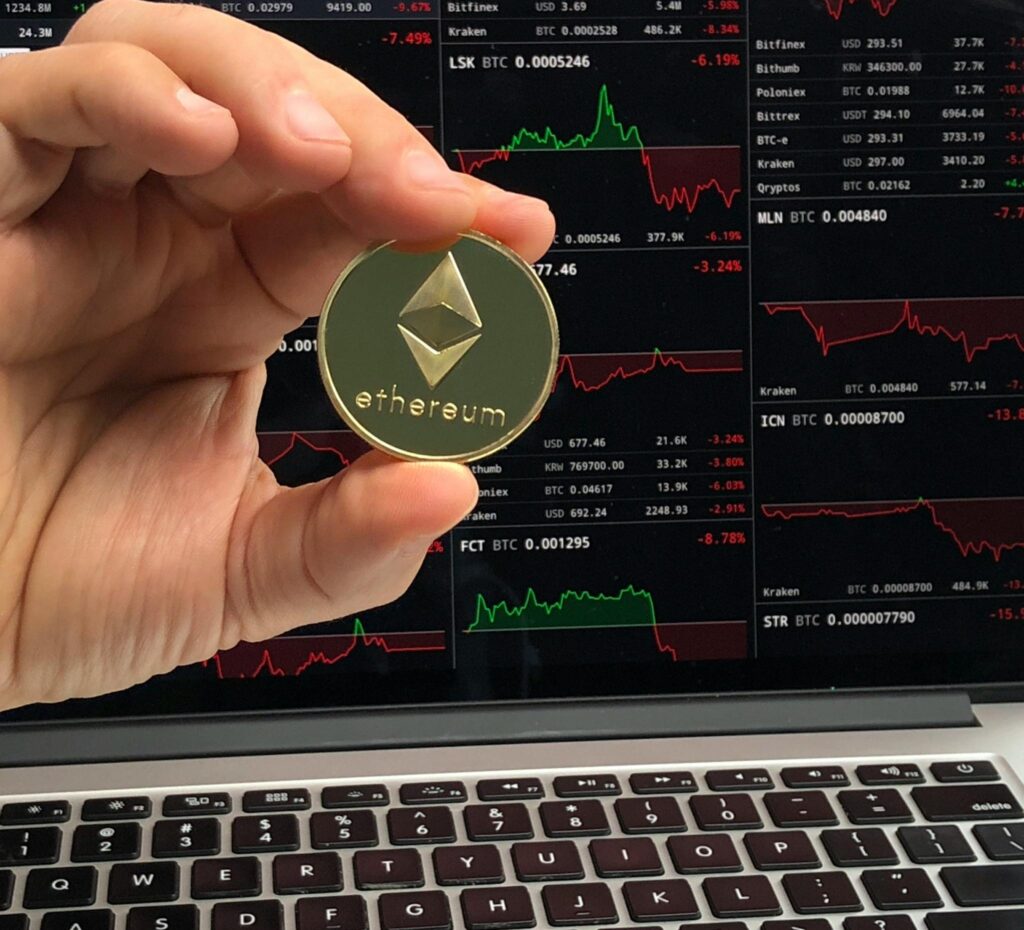 WEB3 - Mundo Crypto A hand holds an Ethereum coin in front of a laptop displaying cryptocurrency trading charts.